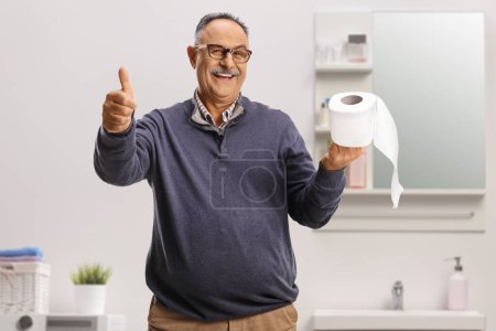 Photo for Smiling mature man holding a toilet paper roll and gesturing thumbs up inside a bathroom - Royalty Free Image