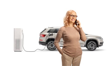 Téléchargez les photos : Femme avec un VUS à un point de charge pour les véhicules électriques faisant un appel téléphonique et souriant isolé sur fond blanc - en image libre de droit