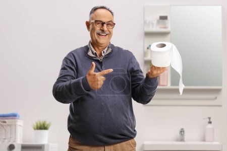 Téléchargez les photos : Souriant homme mûr pointant vers un rouleau de papier toilette à l'intérieur d'une salle de bain - en image libre de droit