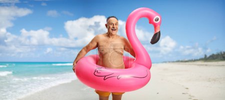 Photo for Happy mature man with a big inflatable flamingo rubber ring posing on a beach - Royalty Free Image
