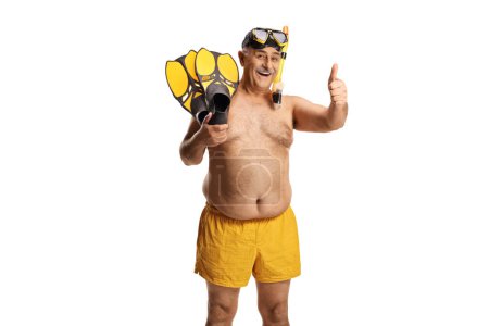 Foto de Hombre maduro en pantalones cortos de natación sosteniendo aletas de snorkel y haciendo gestos con los pulgares hacia arriba aislado sobre fondo blanco - Imagen libre de derechos