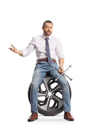 Téléchargez les photos : Homme sérieux assis sur un pneu de voiture et tenant une clé à molette isolée sur fond blanc - en image libre de droit