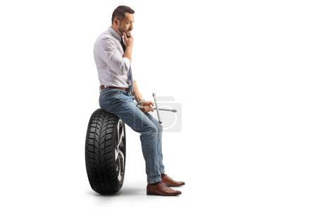 Téléchargez les photos : Photo de profil d'un homme cher assis sur un pneu de voiture et tenant une clé à molette isolée sur fond blanc - en image libre de droit