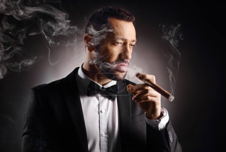 Foto de Joven caballero con traje y pajarita fumando un cigarro sobre un fondo negro - Imagen libre de derechos