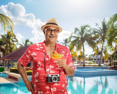 Téléchargez les photos : Touriste heureux avec caméra autour de son cou tenant un cocktail et posant près d'une piscine - en image libre de droit