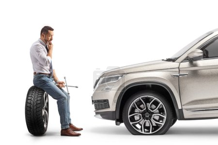 Téléchargez les photos : Photo de profil d'un cher homme assis sur un pneu devant son véhicule et tenant une clé à molette isolée sur fond blanc - en image libre de droit