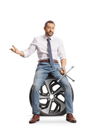 Photo for Serious man sitting on a flat car tire and holding a lug wrench isolated on white background - Royalty Free Image