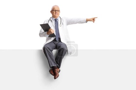 Foto de Médico varón maduro sentado en un panel en blanco y señalando el lado aislado sobre fondo blanco - Imagen libre de derechos