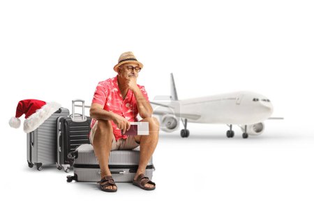 Téléchargez les photos : Toruiste mâle mature assis sur une valise et attendant un vol de Noël retardé devant un avion isolé sur fond blanc - en image libre de droit