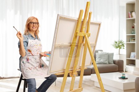 Foto de Mujer sentada en una silla sosteniendo un pincel de pintura y una paleta de colores frente a un lienzo en casa - Imagen libre de derechos