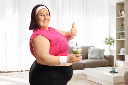 Téléchargez les photos : Femme de taille plus en vêtements de sport tenant un smoothie vert et les pouces geste à la maison - en image libre de droit