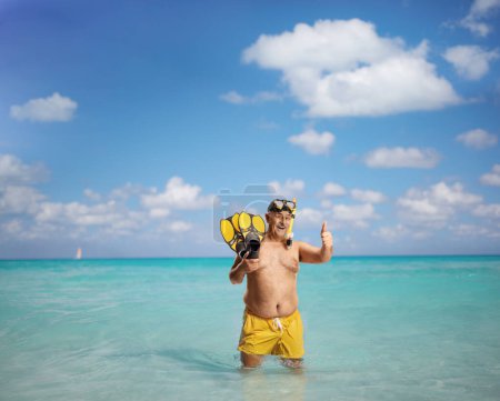 Téléchargez les photos : Homme mûr en short de natation tenant des nageoires de plongée avec tuba et les pouces gestuels dans la mer - en image libre de droit