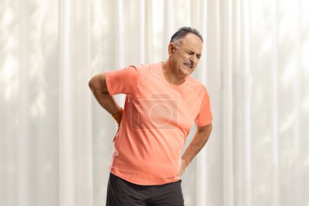 Foto de Hombre maduro en ropa deportiva sosteniendo su columna vertebral en casa - Imagen libre de derechos