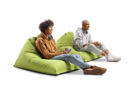 Téléchargez les photos : Africain américain gars et caucasien gars assis sur des sacs de haricots et jouer à un jeu vidéo avec joysticks isolé sur fond blanc - en image libre de droit