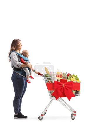 Photo for Full length profile shot of a mother with a baby in a carrier and a shopping cart full of christmas food isolated on white background - Royalty Free Image