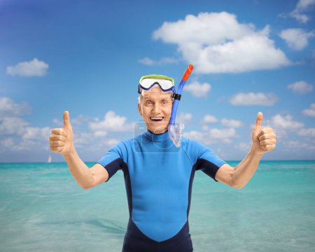Foto de Alegre hombre mayor con equipo de snorkel usando un traje de neopreno y haciendo pulgares hacia arriba signo frente a un mar - Imagen libre de derechos