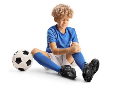 Téléchargez les photos : Garçon avec un ballon de football assis sur le sol et tenant son genou blessé isolé sur fond blanc - en image libre de droit