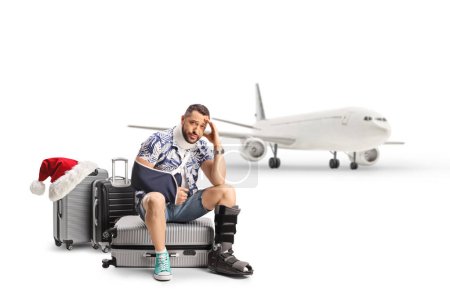 Téléchargez les photos : Homme avec un bras et une jambe blessés assis sur des valises devant un avion, voyageant pour Noël isolé sur fond blanc - en image libre de droit