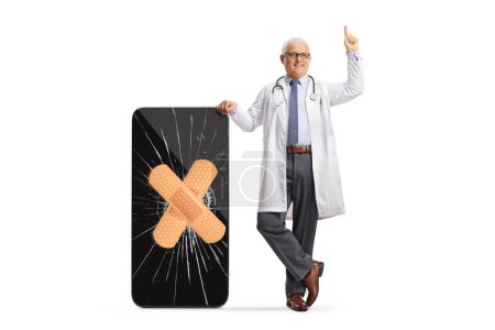 Foto de Médico varón apoyado en un smartphone con una pantalla agrietada y vendaje y apuntando hacia arriba aislado sobre fondo blanco - Imagen libre de derechos
