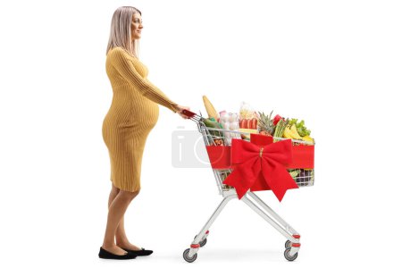 Foto de Foto de perfil de longitud completa de una mujer embarazada empujando un carrito de compras con lazo de cinta roja aislado sobre fondo blanco - Imagen libre de derechos