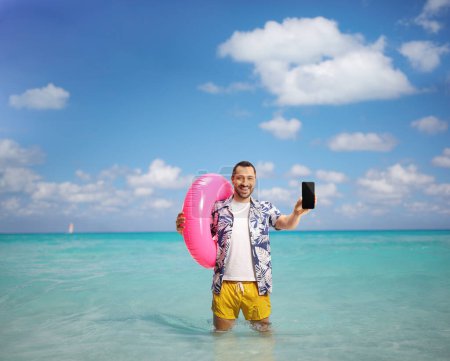Foto de Joven feliz mostrando un teléfono inteligente y sosteniendo un anillo de natación en el mar - Imagen libre de derechos