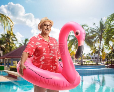 Foto de Turista maduro sonriente con un gran anillo de goma inflable de flamenco junto a una piscina - Imagen libre de derechos