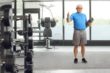 Téléchargez les photos : Portrait complet d'un homme mature en tenue de sport tenant un tapis d'exercice et montrant les pouces dans une salle de gym - en image libre de droit