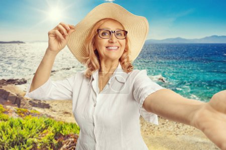 Téléchargez les photos : Belle femme d'âge moyen avec un chapeau de paille prenant un selfie à une plage avec la mer en arrière-plan - en image libre de droit
