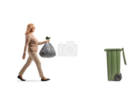 Téléchargez les photos : Photo de profil pleine longueur d'une femme d'âge moyen portant un sac à déchets en plastique et marchant vers un bac isolé sur fond blanc - en image libre de droit