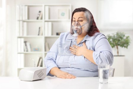 Téléchargez les photos : Femme tenant sa poitrine et utilisant un nébuliseur avec brouillard de vapeur à la maison - en image libre de droit