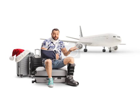 Téléchargez les photos : Touriste masculin avec un bras et une jambe blessés assis sur des valises et attendant un avion, concept de voyage de Noël isolé sur fond blanc - en image libre de droit