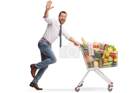 Foto de Hombre alegre corriendo con comida en un carrito de la compra y saludando a la cámara aislado sobre fondo blanco - Imagen libre de derechos