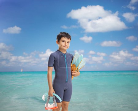 Foto de Niño en traje de neopreno sosteniendo una máscara de buceo y aletas junto al mar - Imagen libre de derechos