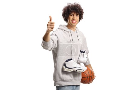 Photo for Smiling young man carrying basketball shoes and a ball and gesturing thumbs up isolated on white background - Royalty Free Image