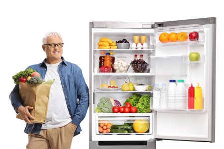 Téléchargez les photos : Homme mûr posant à côté d'un réfrigérateur ouvert et tenant un sac d'épicerie isolé sur fond blanc - en image libre de droit