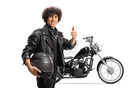 Foto de Joven en una chaqueta de cuero negro haciendo gestos de pulgares delante de una moto aislada sobre fondo blanco - Imagen libre de derechos