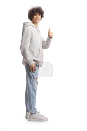 Photo for Full length portrait of a tall guy with curly hair in a gray hoodie and jeans gesturing thumbs up isolated on white background - Royalty Free Image