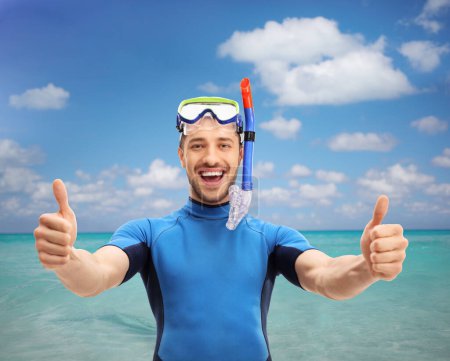 Téléchargez les photos : Guy in a wetsuit with a diving mask making a thumbs up gesture by the sea - en image libre de droit