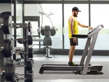 Photo for Full length shot of a bearded guy in sportswear standing on a treadmill and pressing a button at a gym - Royalty Free Image