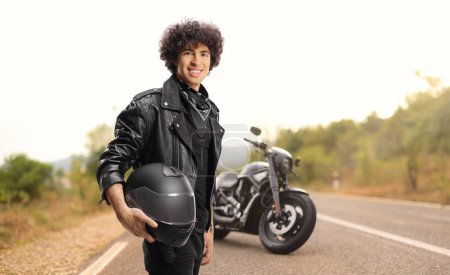 Téléchargez les photos : Young man with curly hair in a black leather jacket holding a helmet in front of a motorbike on the road - en image libre de droit