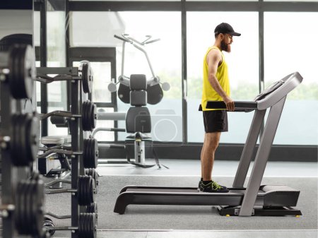 Téléchargez les photos : Bearded guy in sportswear standing on a treadmill and pressing a button at the gym - en image libre de droit