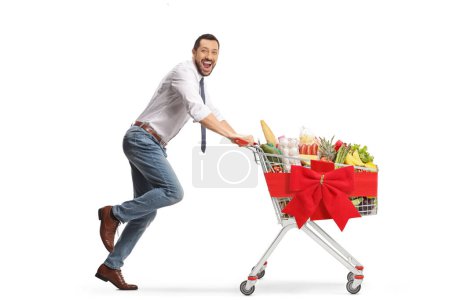 Foto de Excited man running with a shopping cart tied with a bow and smiling at camera isolated on white background - Imagen libre de derechos