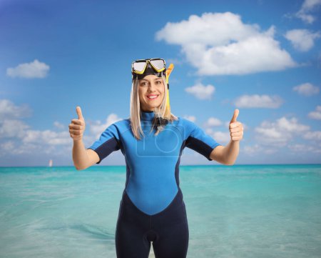 Téléchargez les photos : Young woman in a wetsuit next to the sea with a diving mask and suit gesturing thumbs up - en image libre de droit