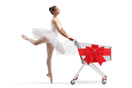 Photo for Full length profile shot of a ballerina in a white tutu dress dancing and pushing a shopping cart tied with red bow isolated on white background - Royalty Free Image