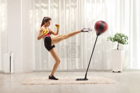 Téléchargez les photos : Jeune femme en forme donnant un coup de pied à un sac de boxe avec la jambe dans une pièce - en image libre de droit