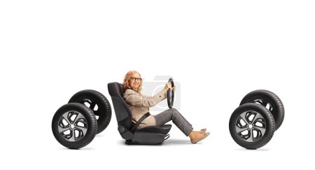 Téléchargez les photos : Femme d'âge moyen assise dans un siège auto sur quatre pneus et tenant un volant isolé sur fond blanc - en image libre de droit
