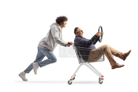 Foto de Joven corriendo y empujando a un hombre maduro con un volante en un carrito de compras aislado sobre fondo blanco - Imagen libre de derechos
