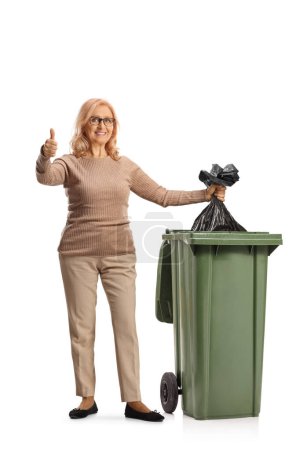 Téléchargez les photos : Femme jetant un sac à déchets en plastique dans une poubelle et geste pouces vers le haut isolé sur fond blanc - en image libre de droit