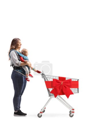 Photo for Full length shot of a mother with a baby in a carrier pushing a shopping cart with red ribbon isolated on white background - Royalty Free Image