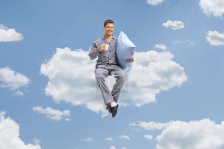 Photo for Young man in pajamas holding a cup and a pillow seated on a cloud up in the sky - Royalty Free Image